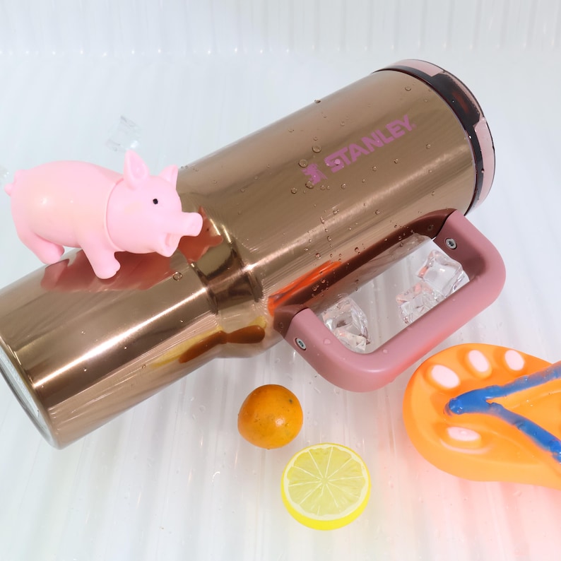 Puede incluir: Un vaso Stanley de color cobre con asa y detalles en rosa, con el logotipo de Stanley. Una peque&ntilde;a figura de cerdo rosa descansa sobre el vaso. Art&iacute;culos adicionales incluyen una naranja peque&ntilde;a, una rodaja de lim&oacute;n y una chancla naranja y azul.