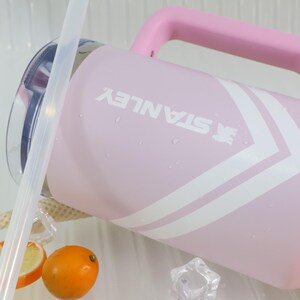 May include: A pink Stanley water jug with a matching handle and white chevron design. The Stanley logo is printed in white. The jug is partially filled with water, and there are ice cubes and a sliced orange in the foreground.