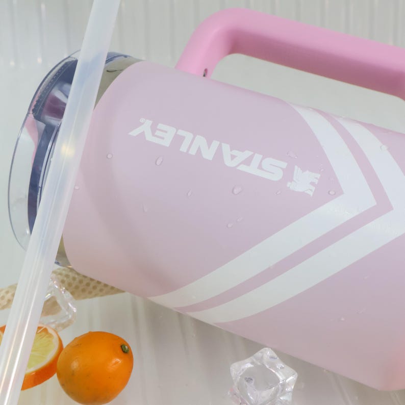 May include: A pink Stanley water jug with a matching handle and white chevron design. The Stanley logo is printed in white. The jug is partially filled with water, and there are ice cubes and a sliced orange in the foreground.