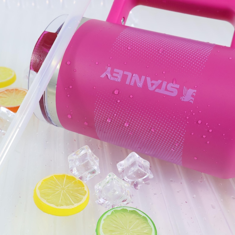 Puede incluir: Una jarra de agua Stanley rosa con asa y el nombre de la marca impreso en el lateral. La jarra est&aacute; parcialmente llena de un l&iacute;quido rojo y tiene una pajita transparente. Cubitos de hielo y rodajas de lim&oacute;n est&aacute;n esparcidos alrededor de la jarra.