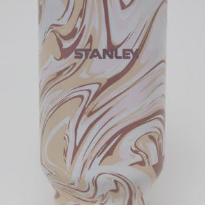 K&ouml;nnte beinhalten: Eine Stanley-Trinkflasche mit einem Wirbelmuster in Wei&szlig;-, Beige- und Braunt&ouml;nen. Das Wort "STANLEY" ist in Dunkelbraun aufgedruckt. Die Flasche hat eine glatte, gl&auml;nzende Oberfl&auml;che.