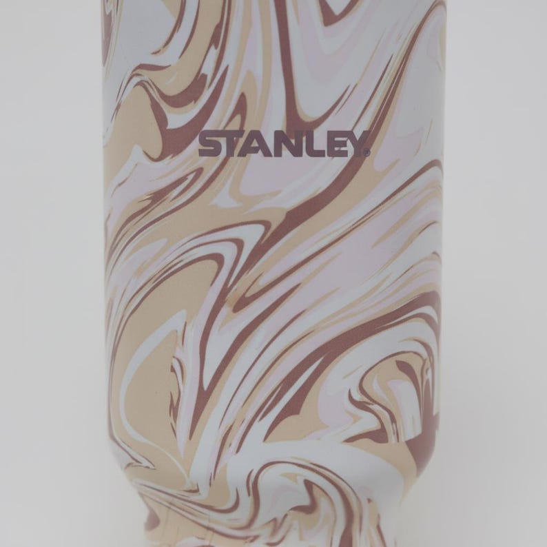 K&ouml;nnte beinhalten: Eine Stanley-Trinkflasche mit einem Wirbelmuster in Wei&szlig;-, Beige- und Braunt&ouml;nen. Das Wort "STANLEY" ist in Dunkelbraun aufgedruckt. Die Flasche hat eine glatte, gl&auml;nzende Oberfl&auml;che.