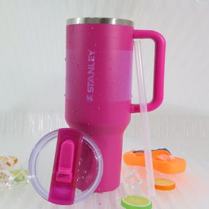 Puede incluir: Un vaso Stanley rosa con asa, tapa y pajita. El vaso tiene el logotipo de Stanley y una banda texturizada. La tapa es transparente con una tapa abatible rosa. Hay una pajita, una chancla naranja y rodajas de fruta.