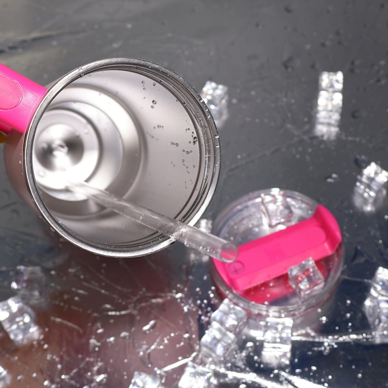 May include: Stainless steel tumbler with a pink handle and lid. The tumbler has a clear straw and is partially filled with water. The lid is clear with a bright pink flip-top. Ice cubes are scattered around the tumbler on a reflective surface.
