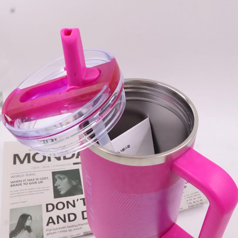 Puede incluir: Un termo de viaje rosa con asa y tapa transparente con pajita rosa. El termo tiene un acabado texturizado y un interior de acero inoxidable. La tapa est&aacute; parcialmente abierta. Un peri&oacute;dico con texto es visible.