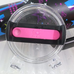 以下が含まれることがあります： 黒い縁と明るいピンクのスライダーが付いた透明なプラスチックタンブラーの蓋。蓋は、カラフルなデザインの黒いタンブラーを部分的に覆っています。デザインには、星、音符、そして「バービー」という言葉が含まれています。
