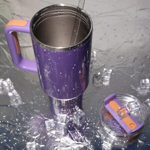 May include: A purple insulated travel mug with a handle and lid. The mug has a stainless steel interior and is covered in water droplets. The handle is purple with a peach-colored accent. Ice cubes are scattered around the mug.