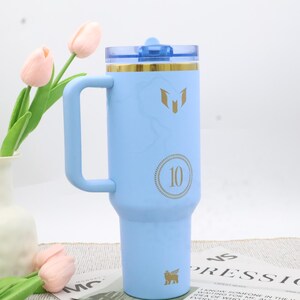 Pu&ograve; includere: Un bicchiere isolante azzurro con manico, coperchio trasparente e dettagli dorati. Il bicchiere presenta un'emblema dorato e il numero "10" in un cerchio dorato. Il bicchiere &egrave; alto circa 23 cm.