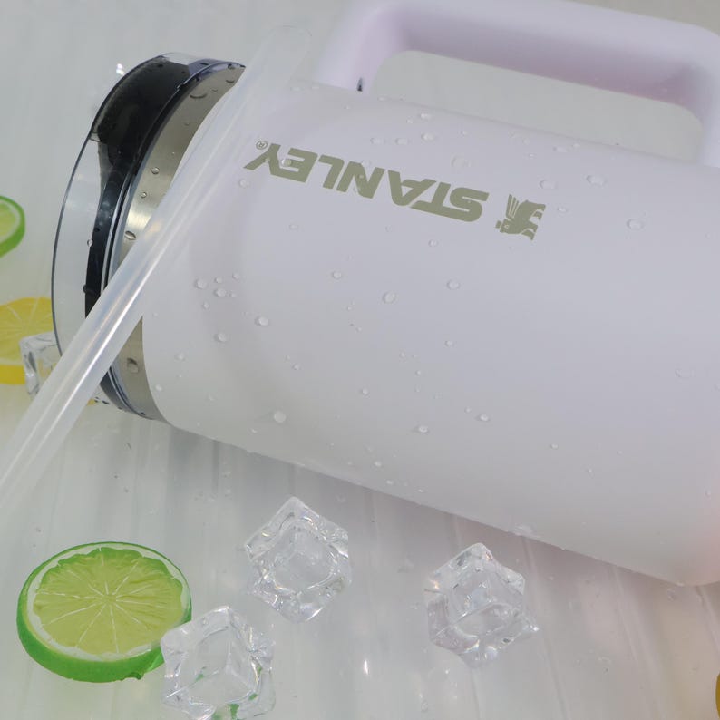 May include: White Stanley water jug with a handle and a clear straw. The jug has the Stanley logo printed on the side. The jug is wet with water droplets and surrounded by ice cubes and a lime slice.