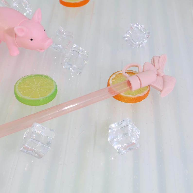 K&ouml;nnte beinhalten: Ein rosa Strohhalm in Schweineform mit Schleife, der auf einer Orangenscheibe liegt. Das Bild enth&auml;lt auch eine Limettenscheibe, Eisw&uuml;rfel und einen rosa Strohhalm. Die Artikel sind auf einer wei&szlig;en Oberfl&auml;che angeordnet.