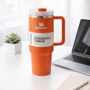 May include: Orange Stanley Quencher H2.0 Tumbler with a handle, straw, and white lid. The tumbler has the Stanley logo and product information printed on the side. The tumbler is on a white surface next to a laptop and notebook.