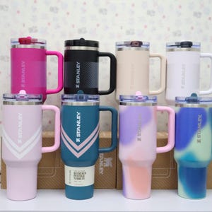 Puede incluir: Una colecci&oacute;n de vasos Stanley Quencher en varios colores, incluyendo rosa, negro, blanco y turquesa. Cada vaso tiene un asa y una tapa. El logotipo de Stanley es visible en el lateral.