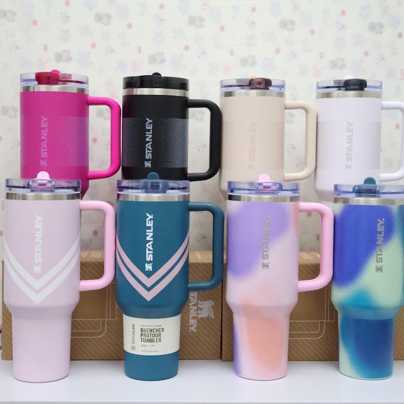 Puede incluir: Una colecci&oacute;n de vasos Stanley Quencher en varios colores, incluyendo rosa, negro, blanco y turquesa. Cada vaso tiene un asa y una tapa. El logotipo de Stanley es visible en el lateral.