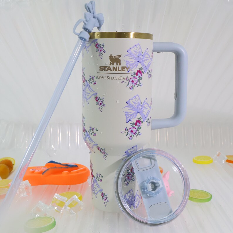 May include: A white Stanley tumbler with a floral and bow pattern, a light blue handle, and a matching straw. The tumbler has a gold rim and the text "STANLEY" and "LOVESHACKFANCY". The lid is clear with a light blue slider.