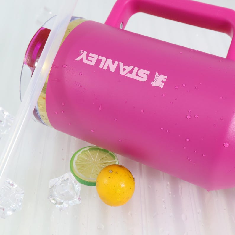 May include: A bright pink Stanley water jug with a handle and the brand name in white. The jug is next to a lemon slice, a whole lemon, ice cubes, and a clear straw.