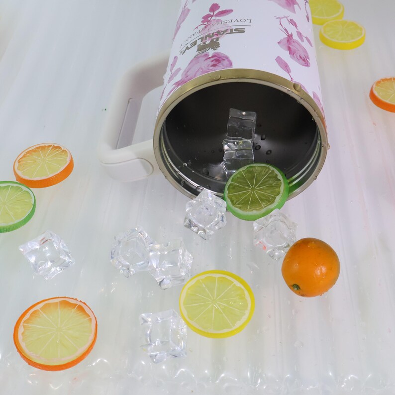 Pode incluir: Um copo Stanley branco com estampa floral rosa, com al&ccedil;a, cheio de cubos de gelo e uma fatia de lim&atilde;o. Espalhadas, fatias de laranja, lim&atilde;o e lim&atilde;o, e mais cubos de gelo. O copo tem uma borda dourada.