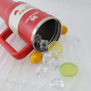 Puede incluir: Un vaso t&eacute;rmico rojo Stanley H2.0 con asa, lleno de cubitos de hielo, rodajas de lim&oacute;n y lima, y naranjas peque&ntilde;as. El vaso tiene una etiqueta blanca con el nombre de la marca y un s&iacute;mbolo de coraz&oacute;n.