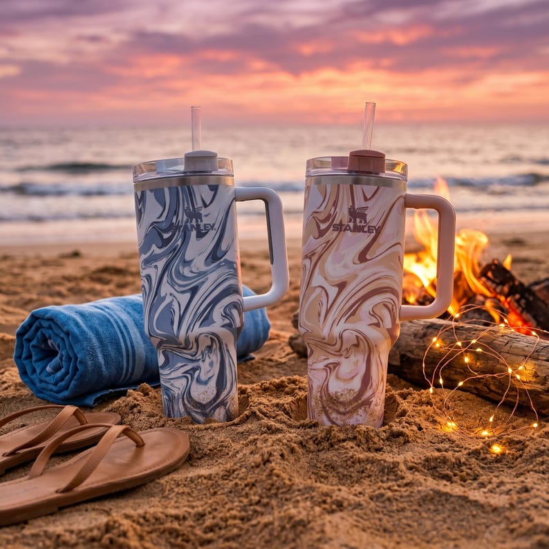 May include: Two large insulated tumblers with handles and straws, one in a blue and white swirl pattern, the other in a brown and white swirl pattern. The tumblers are on a sandy beach with a blue towel and sandals.