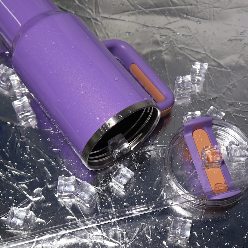 May include: A purple insulated water bottle with a handle and a clear lid. The bottle has a silver rim and a peach-colored accent on the handle and lid. Ice cubes are scattered around the bottle on a reflective surface.