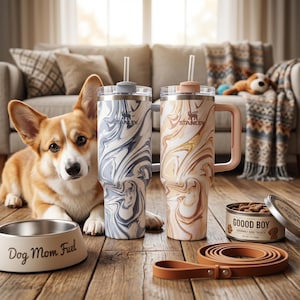 May include: A corgi dog lies next to a dog bowl that reads "Dog Mom Fuel". Two Stanley tumblers with marble designs and straws sit next to the dog. A can of dog treats and a leather leash are also in the image.