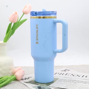 Pu&ograve; includere: Un bicchiere termico Stanley azzurro con manico e coperchio blu. Il bicchiere ha un bordo color oro e il logo Stanley in oro. Il bicchiere &egrave; cilindrico con una base pi&ugrave; ampia e una parte superiore pi&ugrave; stretta.