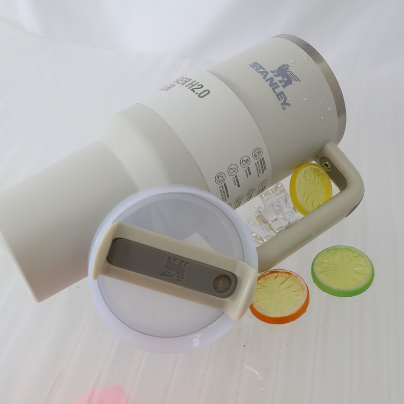 May include: A white Stanley H2.0 tumbler with a handle and a clear lid. The tumbler has the Stanley logo and product information printed on it. Decorative lemon and lime slices are placed near the tumbler.