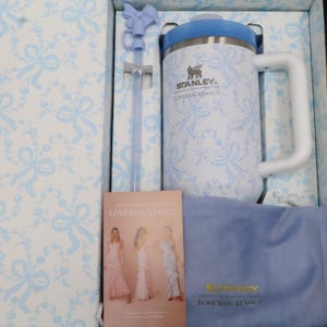 K&ouml;nnte beinhalten: Ein wei&szlig;-blauer Stanley Loveshackfancy-Becher mit gro&szlig;em Griff, blauem Deckel und passendem Strohhalm. Der Becher befindet sich in einer Schachtel mit passendem Design und einem kleinen Booklet. Ein blaues Tuch ist ebenfalls enthalten.