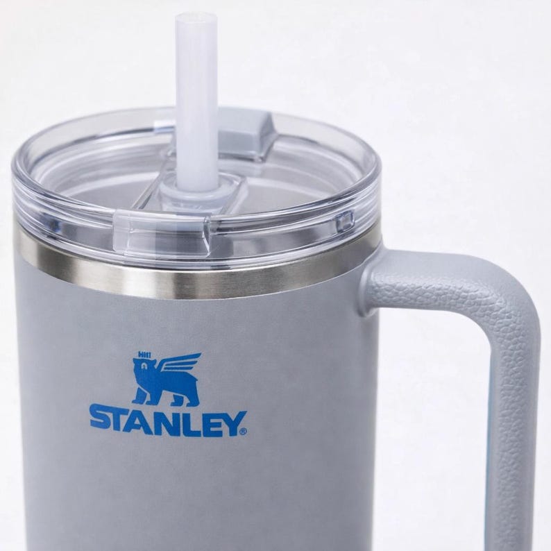 May include: A gray Stanley insulated tumbler with a clear lid and white straw. The tumbler has a handle and the Stanley logo in blue. The tumbler is designed to keep drinks cold or hot.
