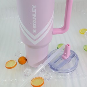 Puede incluir: Un vaso Stanley rosa con asa y dise&ntilde;o de galones blancos. El vaso tiene tapa transparente, pajita y est&aacute; rodeado de cubitos de hielo y rodajas de fruta. El logotipo de Stanley es visible.