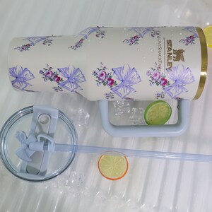 May include: A cream-colored Stanley tumbler with a floral pattern of purple bows and pink roses. The tumbler has a light blue handle and a clear lid with a straw. The tumbler is decorated with the text "LOVESHACKFANCY".