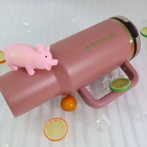 May include: A dusty rose Stanley tumbler with a handle, the Stanley logo in green, and a pink pig figurine. The tumbler is surrounded by ice cubes and citrus fruit slices.