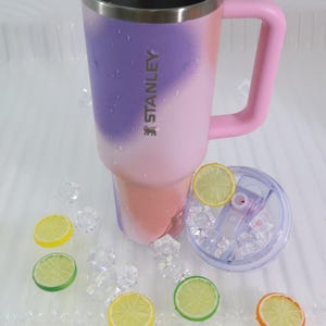 Puede incluir: Un vaso t&eacute;rmico Stanley con asa rosa y dise&ntilde;o degradado en rosa, morado y coral. El vaso tiene el logotipo de Stanley y se muestra con una tapa transparente, hielo y rodajas de lim&oacute;n.