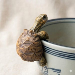 Może przedstawiać: Szczegółowa, brązowa figurka żółwia trzymająca się krawędzi białej i niebieskiej ceramicznej miski. Żółw ma teksturowany pancerz i szczegółowe cechy. Miska ma niebieski wzór w paski.