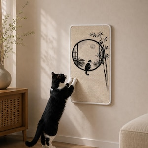 Op de afbeelding: Een rechthoekige krabplank voor aan de muur met een beige textuuroppervlak, voorzien van een zwarte inktillustratie van een rond raam, bamboestengels en het silhouet van een kat die op het kozijn zit, afgewerkt met een witte rand.