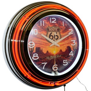 May include: A round wall clock featuring a bright orange neon ring and a glass face. The clock dial displays a desert landscape at sunset with a Route 66 shield logo, the text The Mother Road, and white numbers from 1 to 12 on a dark background.
