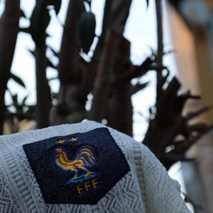 May include: A white, open-weave sweater with a navy blue patch displaying a gold and blue rooster emblem, two gold stars, and the letters "FFF". The sweater is shown outside with a blurred tree backdrop.