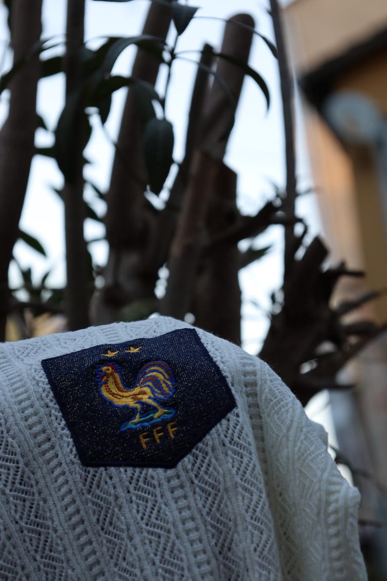 May include: A white, open-weave sweater with a navy blue patch displaying a gold and blue rooster emblem, two gold stars, and the letters "FFF". The sweater is shown outside with a blurred tree backdrop.