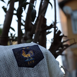 May include: A cream-coloured, open-knit sweater with a navy blue patch displaying a gold rooster emblem and the letters "FFF". The sweater has a diamond pattern. The background includes tree branches and a building.