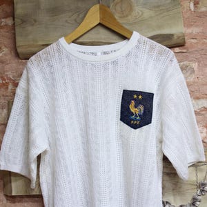 May include: White short-sleeved shirt with a textured knit pattern. The shirt features a navy blue pocket with a gold rooster emblem and the letters "FFF". The shirt is hanging on a wooden hanger.