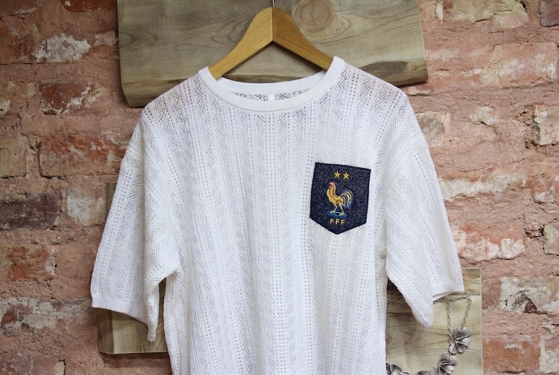 May include: White short-sleeved shirt with a textured knit pattern. The shirt features a navy blue pocket with a gold rooster emblem and the letters "FFF". The shirt is hanging on a wooden hanger.