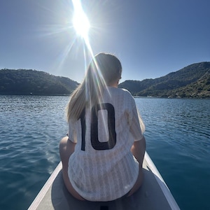 May include: A person wearing a white jersey with the number 10 on the back, sitting on a paddleboard on a lake. The sun shines brightly in the background, with mountains and water visible.