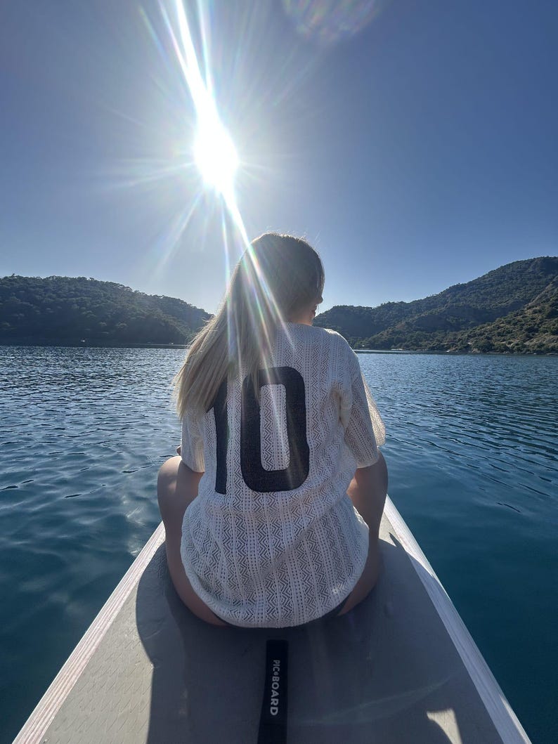 May include: A person wearing a white jersey with the number 10 on the back, sitting on a paddleboard on a lake. The sun shines brightly in the background, with mountains and water visible.