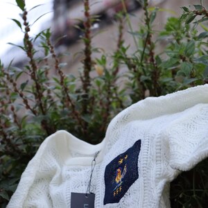 May include: A white, open-weave sweater with a crew neck and short sleeves. The sweater features a navy blue patch with a gold rooster and the letters "FFF". A tag with the brand name "LISCIO" hangs from the sweater.