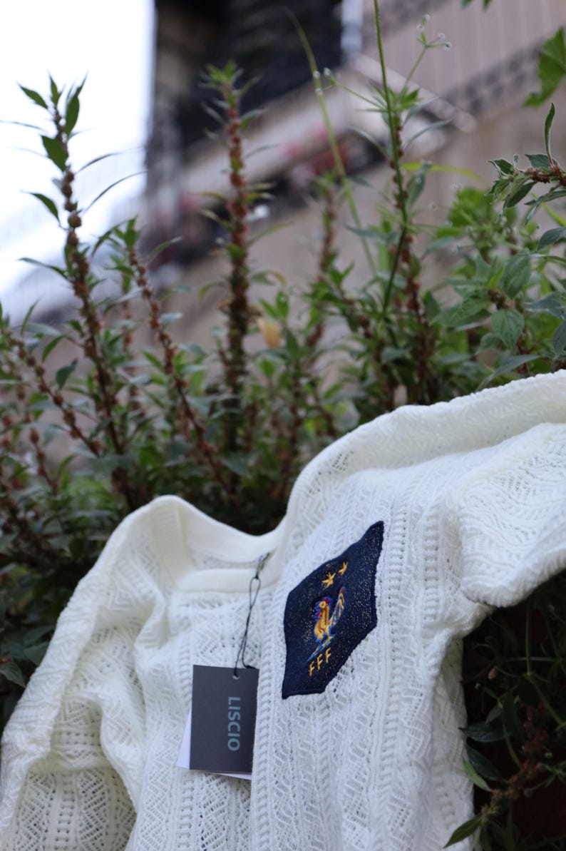 May include: A white, open-weave sweater with a crew neck and short sleeves. The sweater features a navy blue patch with a gold rooster and the letters "FFF". A tag with the brand name "LISCIO" hangs from the sweater.