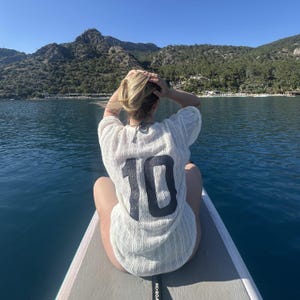 May include: A person wearing a white, open-weave shirt with the number 10 in black on the back, sitting on a paddleboard. The background shows water, mountains, and a clear blue sky.