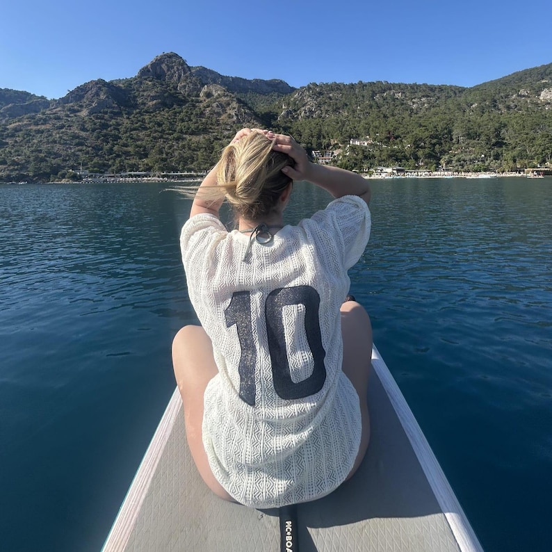 May include: A person wearing a white, open-weave shirt with the number 10 in black on the back, sitting on a paddleboard. The background shows water, mountains, and a clear blue sky.