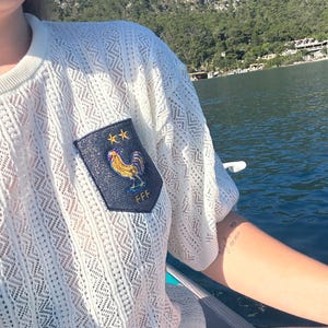 May include: Off-white, open-weave knit top with short sleeves. A navy blue patch on the chest displays a gold rooster, two gold stars, and the letters "FFF". The top is worn outdoors near water.