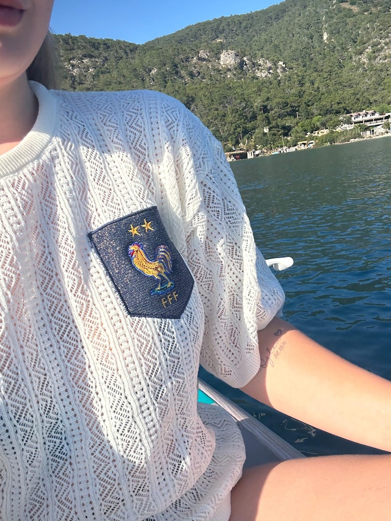 May include: Off-white, open-weave knit top with short sleeves. A navy blue patch on the chest displays a gold rooster, two gold stars, and the letters "FFF". The top is worn outdoors near water.