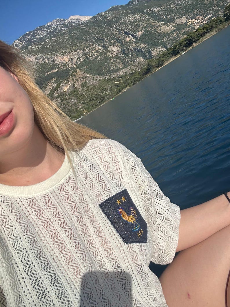 May include: A cream-coloured, open-weave short-sleeved shirt with a crew neck. A navy blue patch on the shirt features a golden rooster, two stars, and the letters "FFF". The background shows a lake and mountains under a bright blue sky.