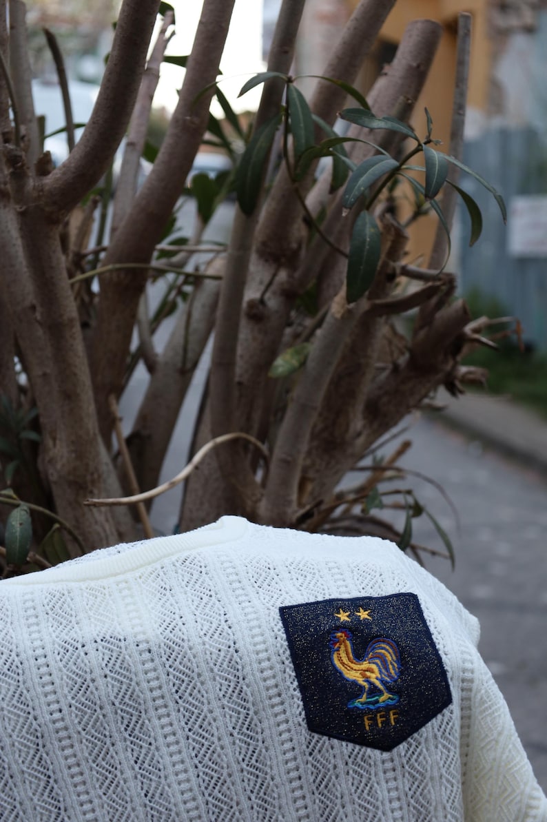 May include: Off-white open-knit sweater with a navy patch displaying a gold rooster emblem and the letters "FFF". The sweater has a crew neck and a textured, diamond-patterned knit. The background features tree branches and a blurred street scene.